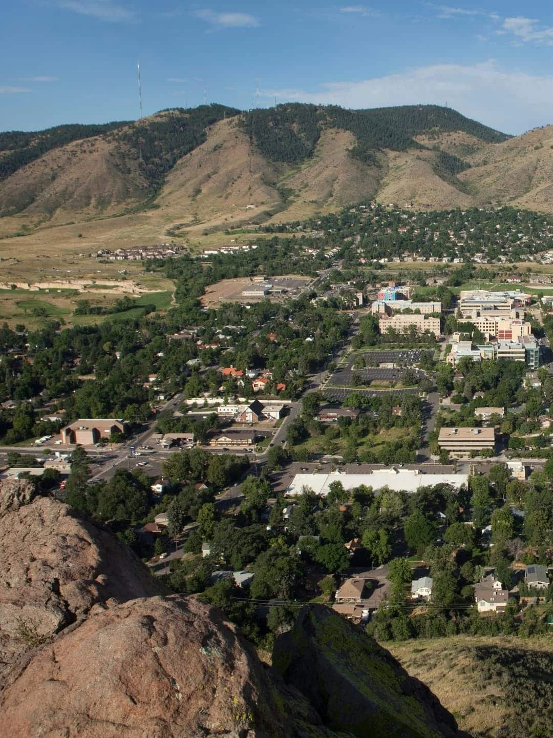 Aerial view of a city with a mix of residential and commercial buildings, surrounded by green trees and mountains in the background under a blue sky.
