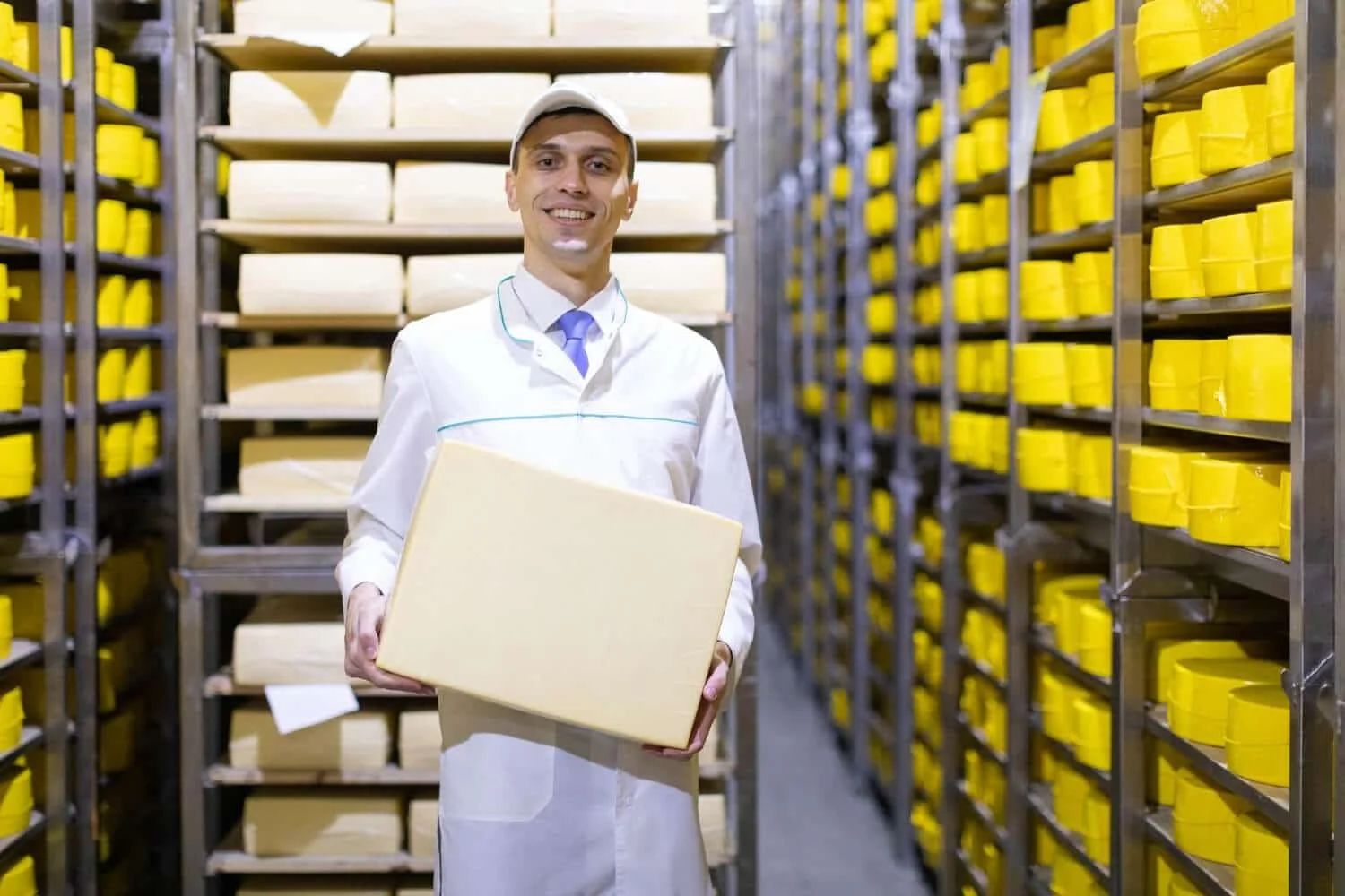 A man in a white coat holding a box in a cheese storage room with shelves of cheese wheels and blocks.