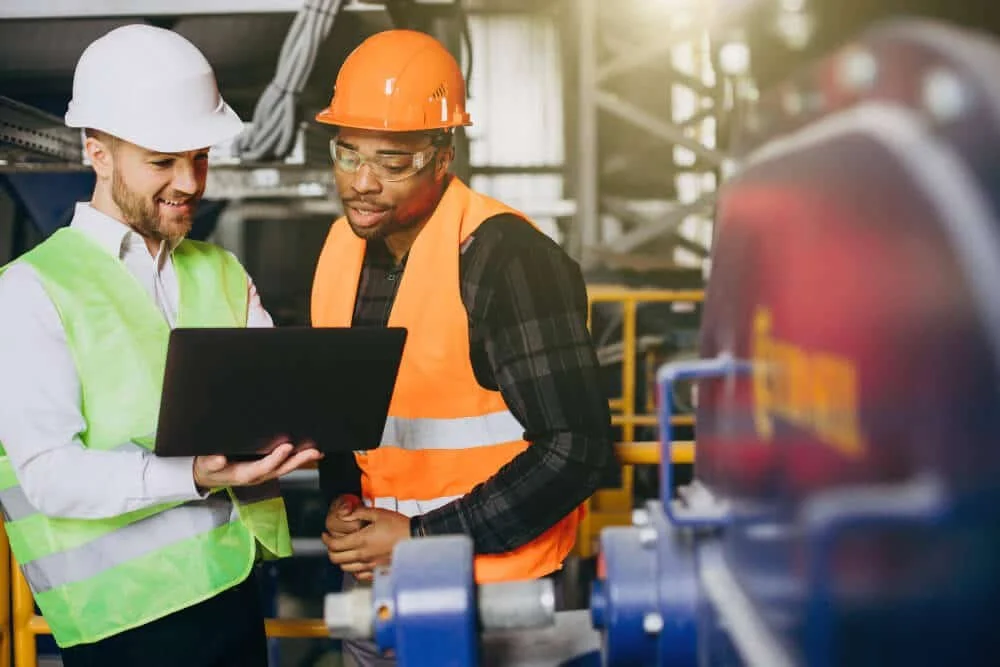 Two male factory workers wearing safety helmets and reflective vests looking at a laptop in an industrial setting.