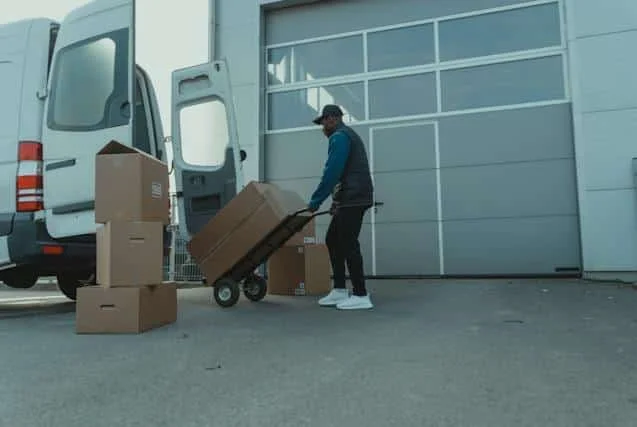 A man loading boxes onto a hand truck outside a delivery van in front of a large garage door.