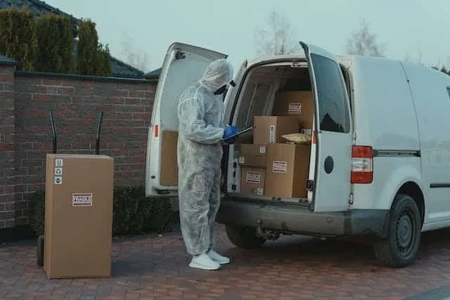Person in protective suit unloading boxes from a white cargo van outdoors.