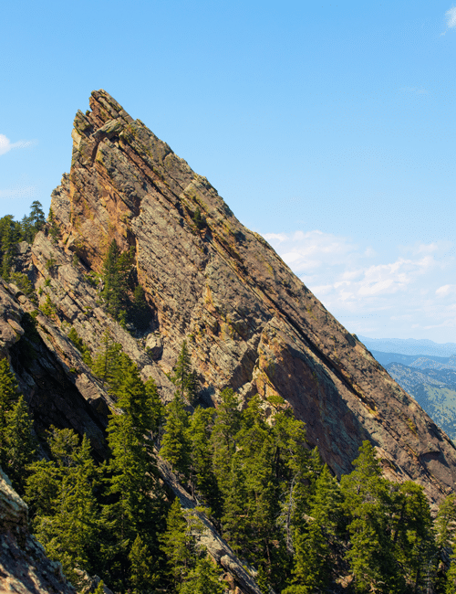 A sharp, jagged mountain peak with vertical rock formations and surrounding green pine trees under a blue sky.