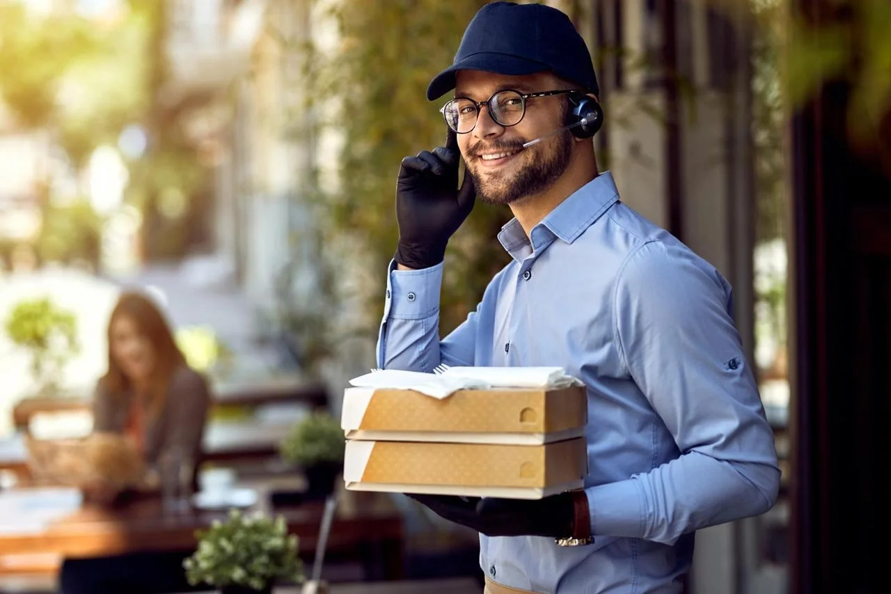 A man wearing glasses, a blue shirt, and a black hat is smiling and talking on a headset. He is holding three pizza boxes and has an open pizza box on top. There is a woman sitting at a table in the background of the outdoor patio.