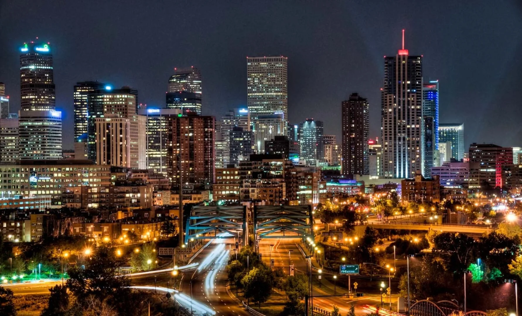 Night Denver skyline with illuminated skyscrapers, city streets with light trails from moving vehicles, and a dark sky overhead.