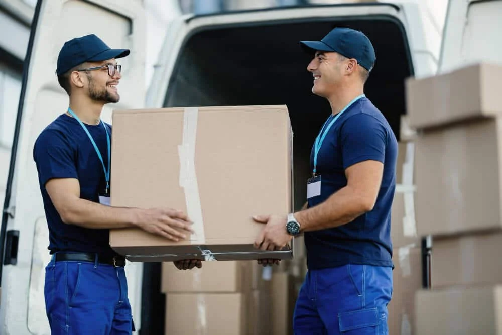 Two delivery workers in blue uniforms and hats exchanging a cardboard box in front of a delivery van.