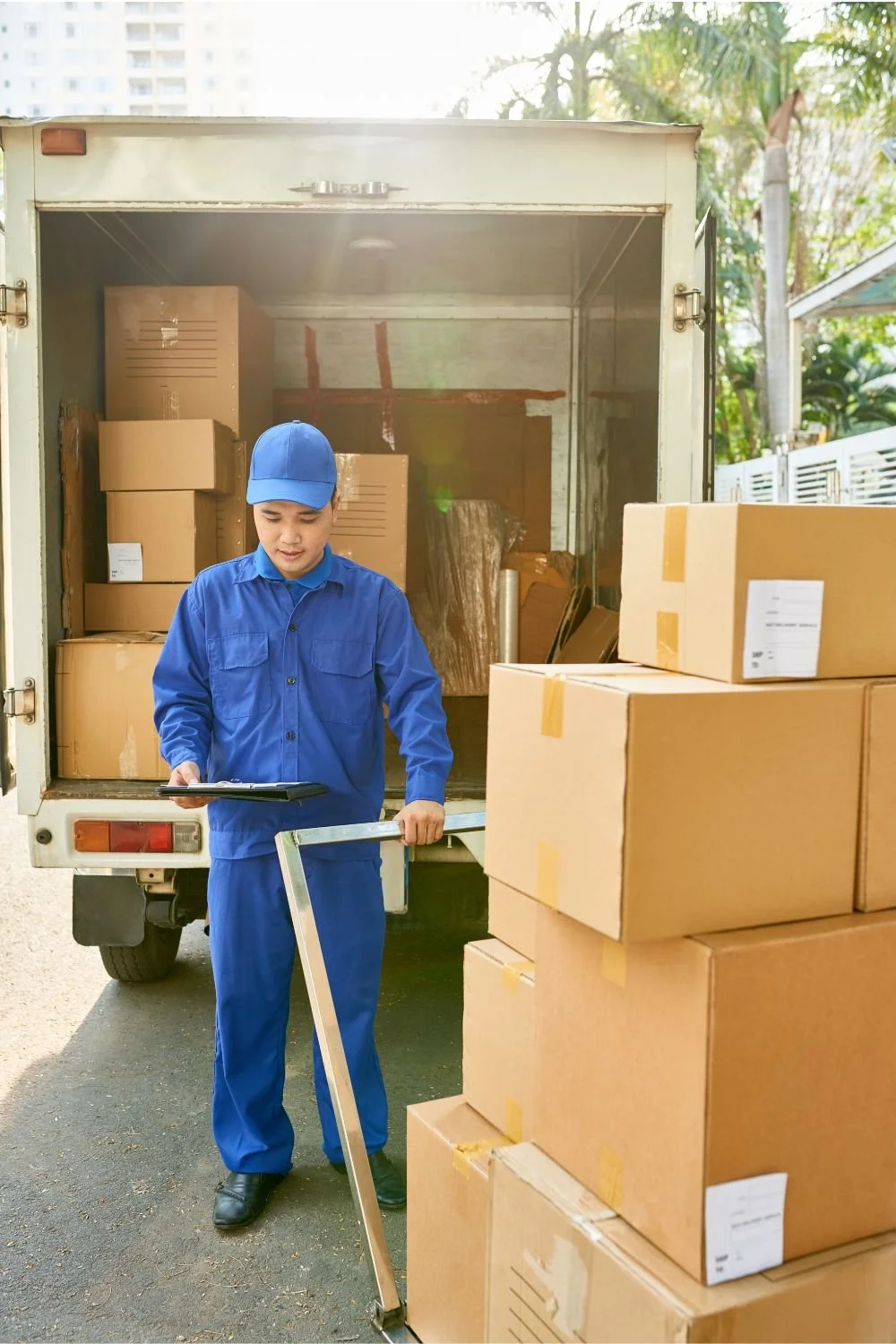 A delivery person in blue uniform standing outside his delivery truck, holding a clipboard and a hand truck. Several cardboard boxes are stacked beside him and inside the truck.