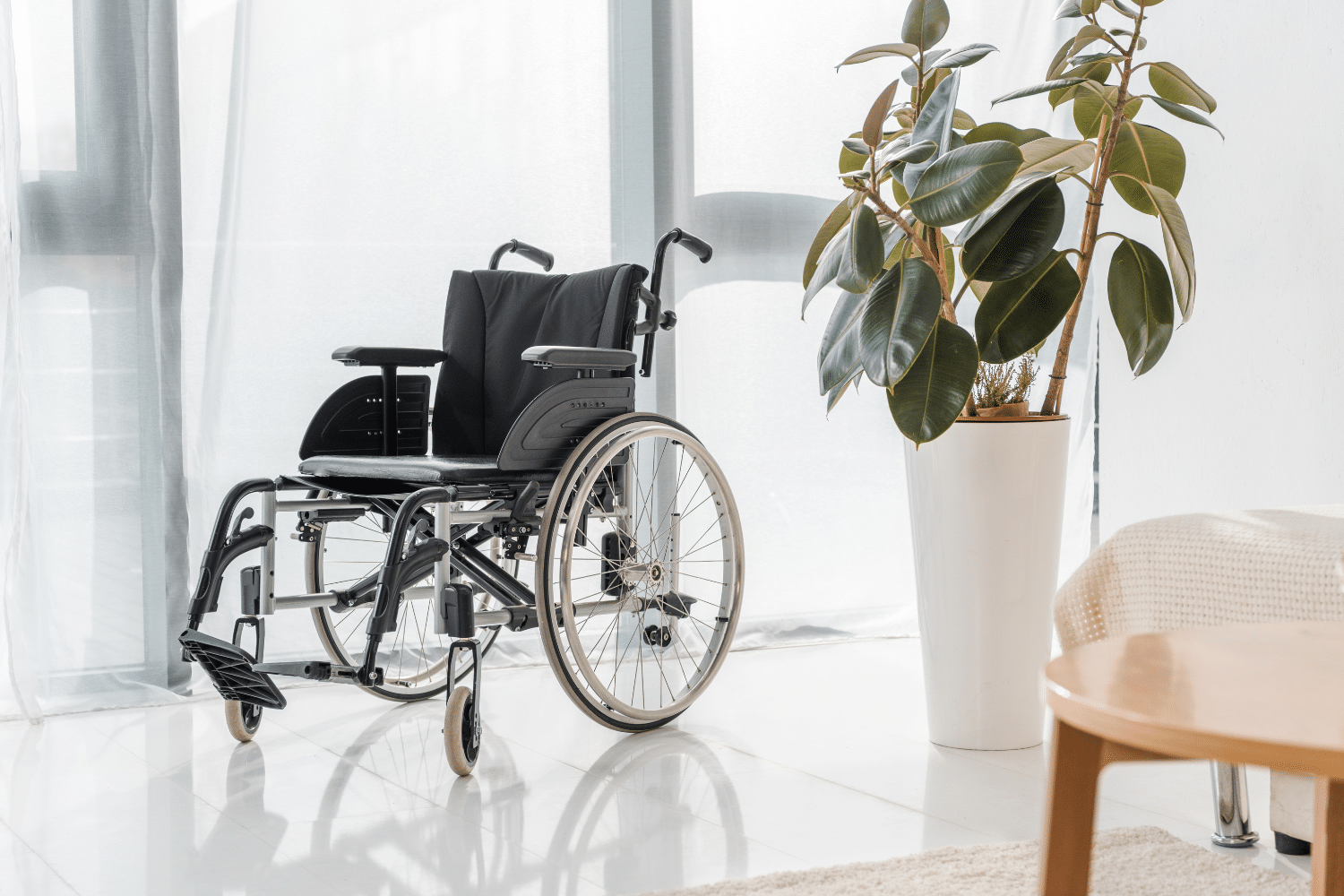 Wheelchair next to tall potted plant in a room with white curtains and floor, beige chair, and wooden table.