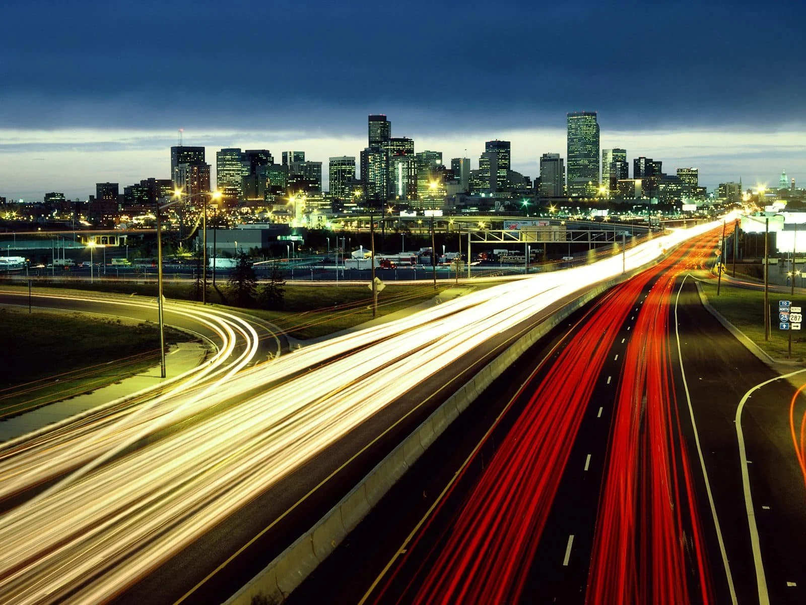 Nighttime cityscape of Denver with light trails from moving vehicles on a busy highway leading to a city skyline with tall buildings, under a cloudy sky.