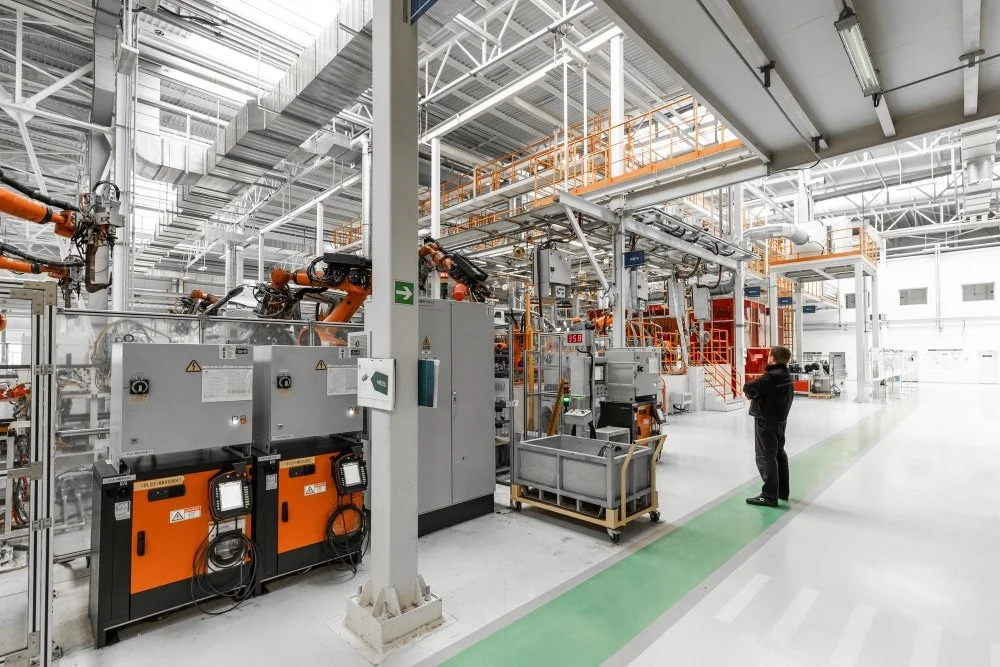A person in a black jacket and pants standing in an industrial factory with large machinery, robotic arms, and metal piping, illuminated by bright overhead lighting.