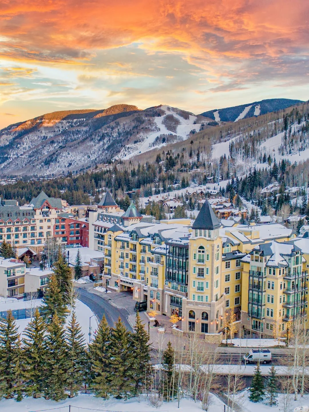 A mountain town with colorful buildings at sunset, snow-covered slopes and trees in the background.