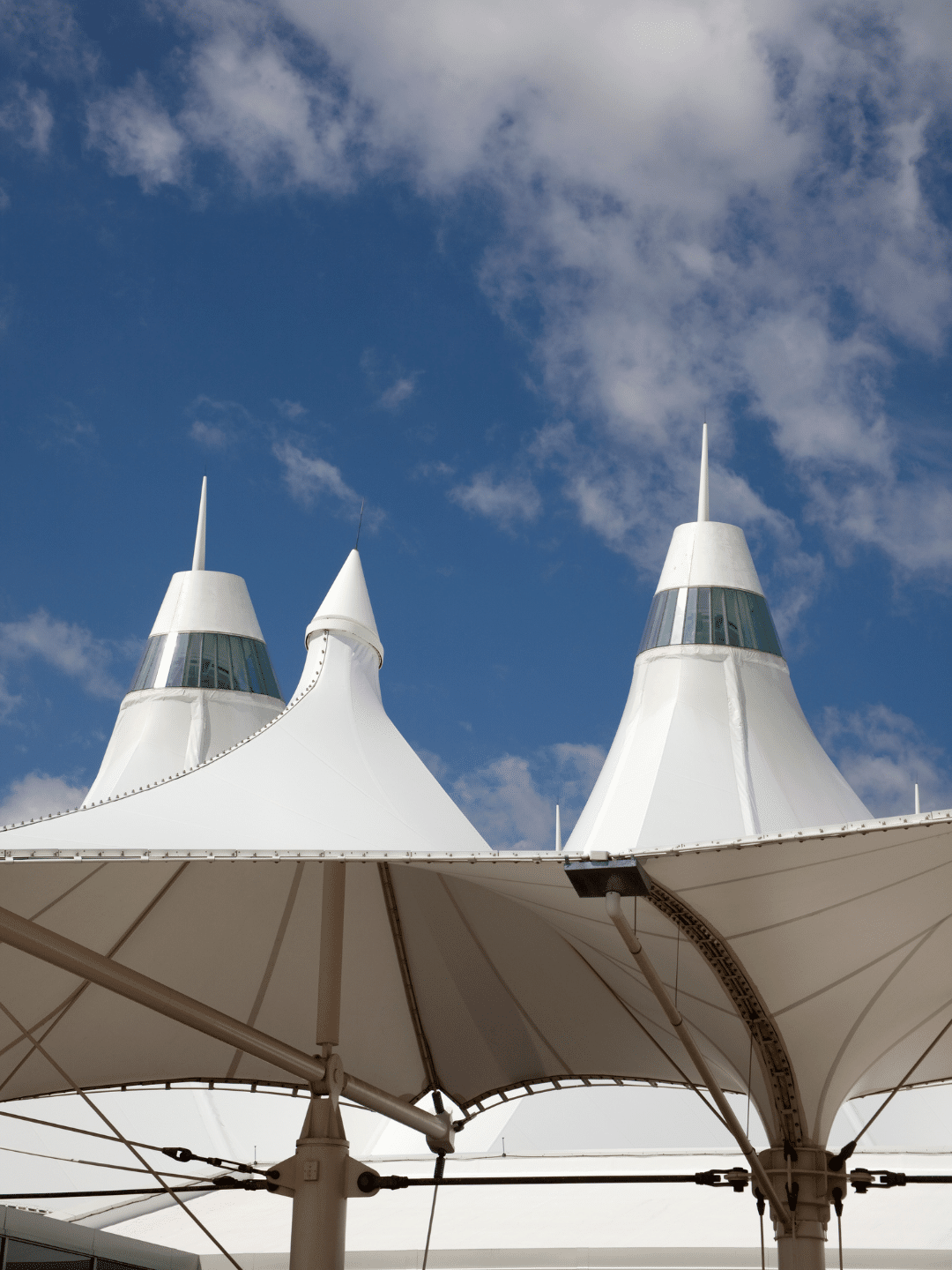 Modern architectural structure with white, tent-like fabric roofs and pointed towers against a partly cloudy blue sky.