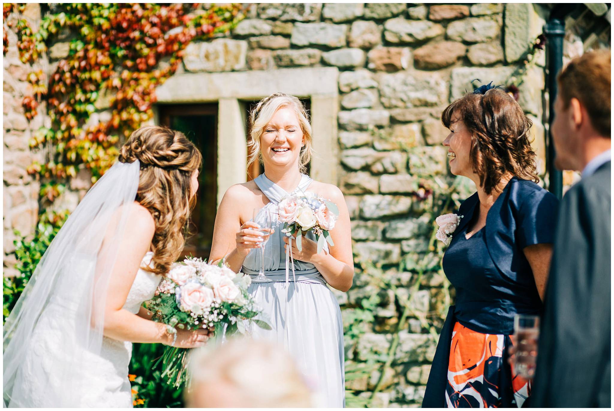 bridesmaid laughing with eyes closed holding glass of chapagne and flowers outside browsholme hall and tithe barn