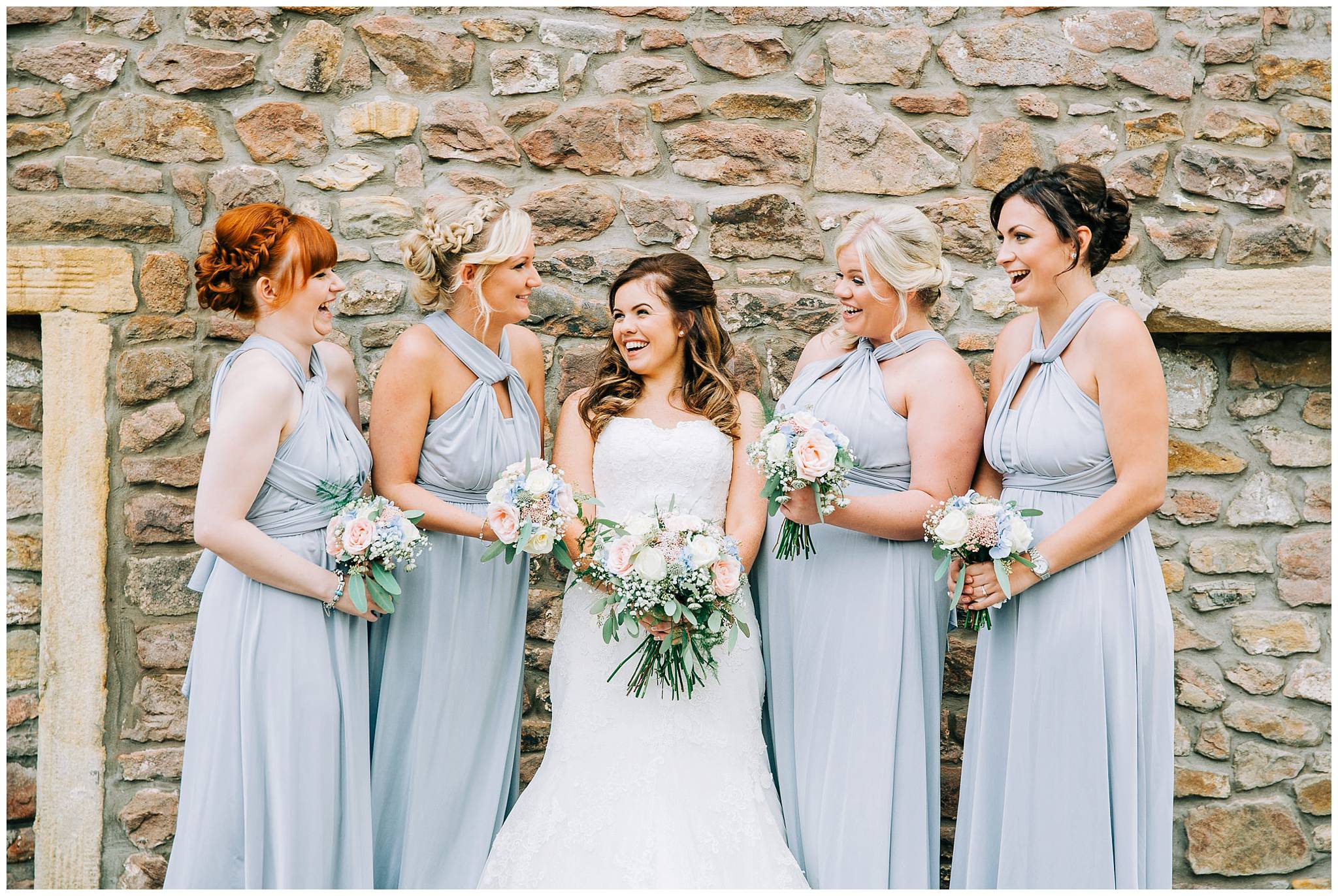 bridesmaids posed smiling at each other against browsholme hall tithe barn stone wall