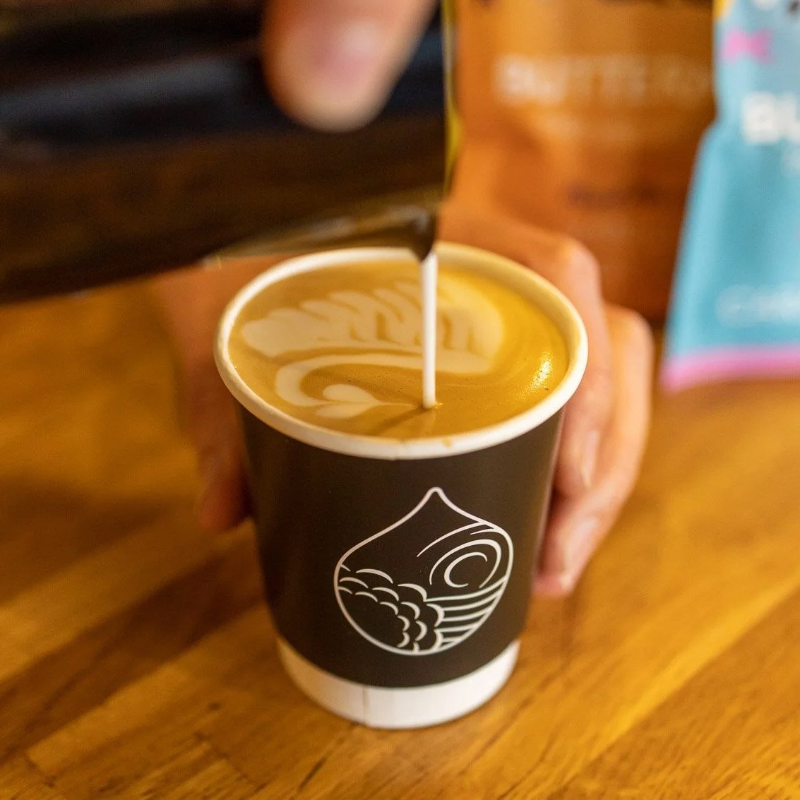 A barista artfully pours milk into a black recycled takeaway coffee cup with the Bean Shot Droplet Mnemonic.