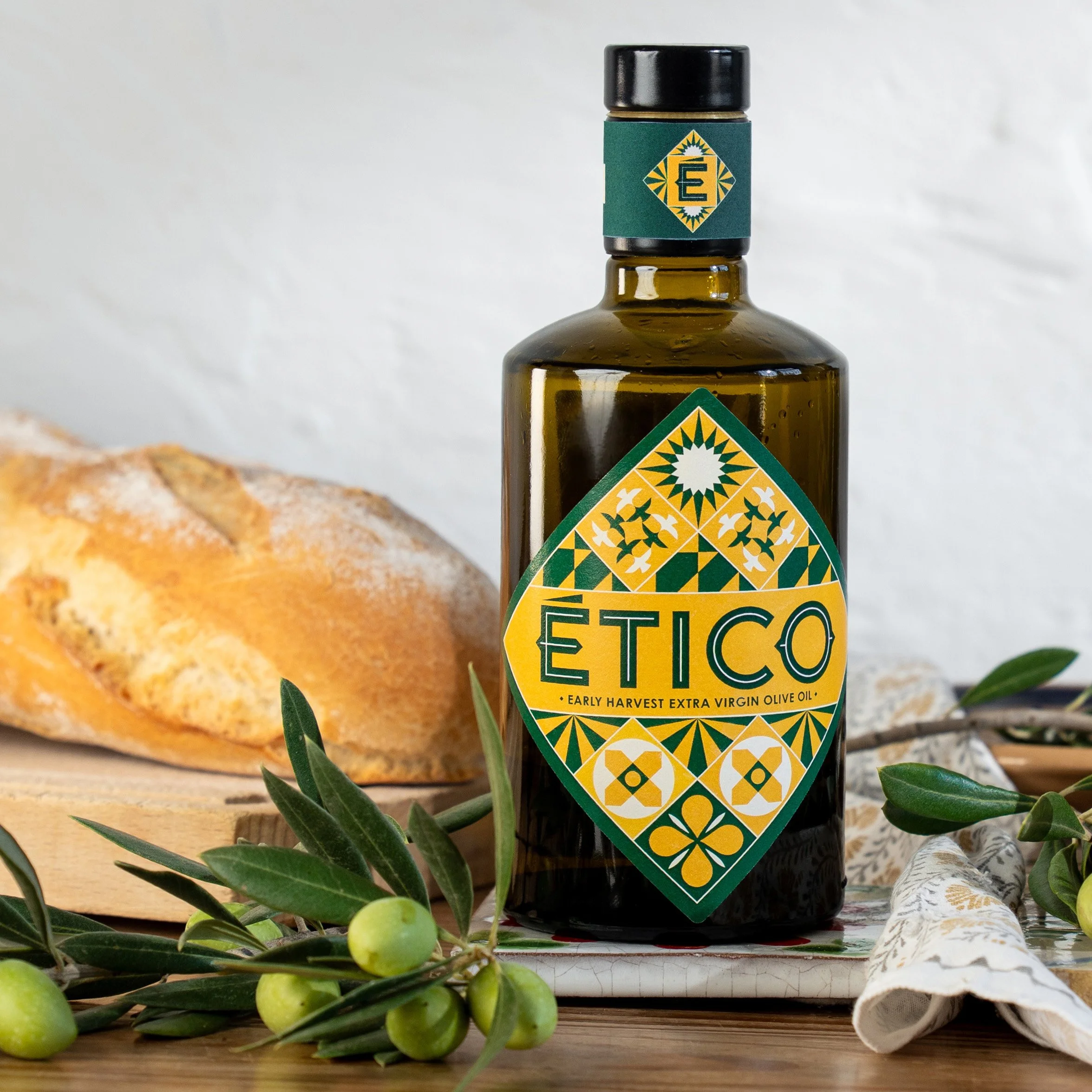 A bottle of Ético Extra Virgin Olive Oil sat with fresh bread and olives. The label features Moorish inspired tile patterns that tell the ethical story of the oil and it’s production.