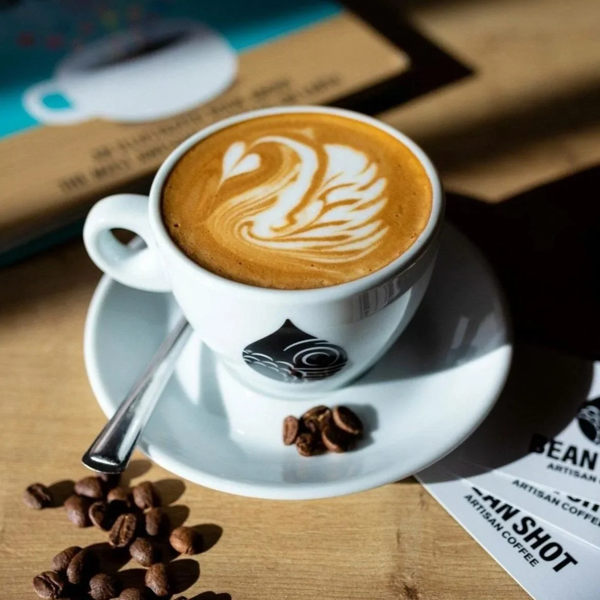 A Bean Shot Coffee cup and saucer with a fresh cappuccino featuring swan coffee art. The cup is propped with fresh coffee beans.