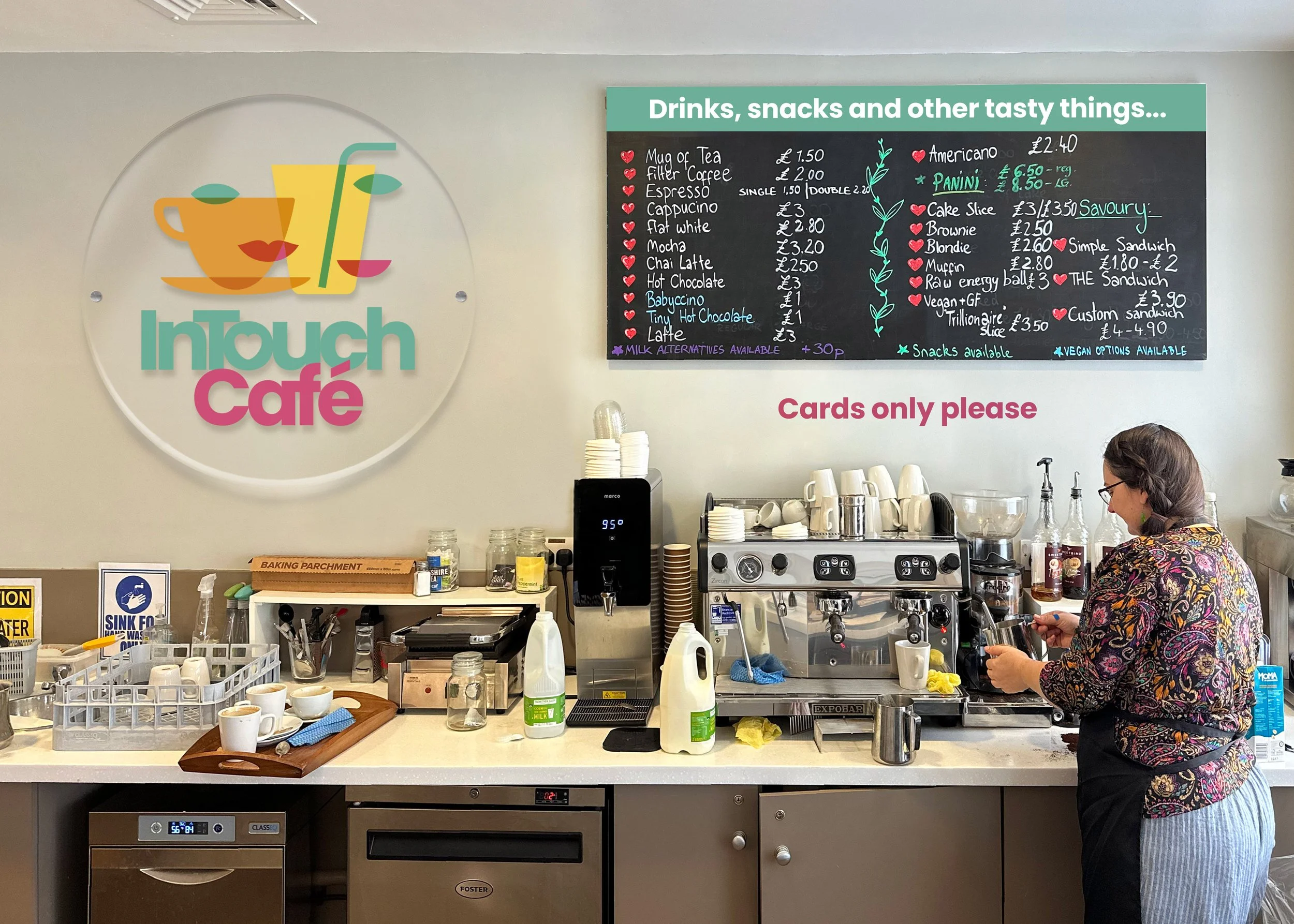Interior of InTouch Café counter with a colourful logo that says "InTouch Café" on the wall, a woman preparing a beverage, a blackboard menu listing drinks and snacks, and various coffee-making equipment and supplies.
