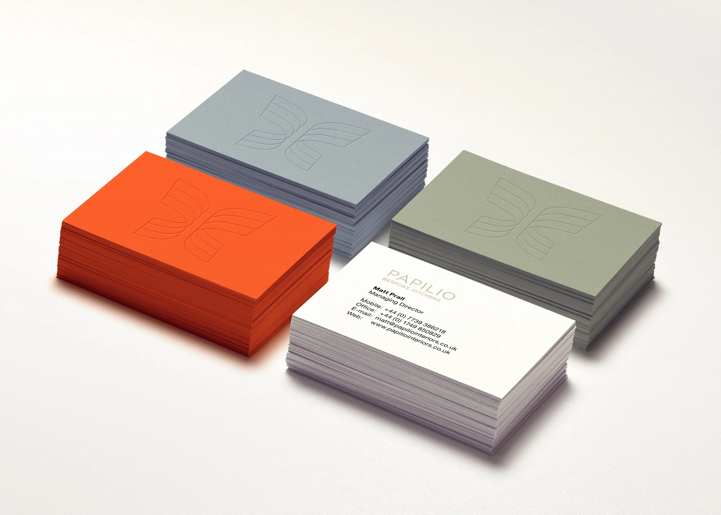 Stacks of business cards in orange, gray, and green colors on a white surface, with one set displaying contact information for Papilio Bespoke Kitchens.