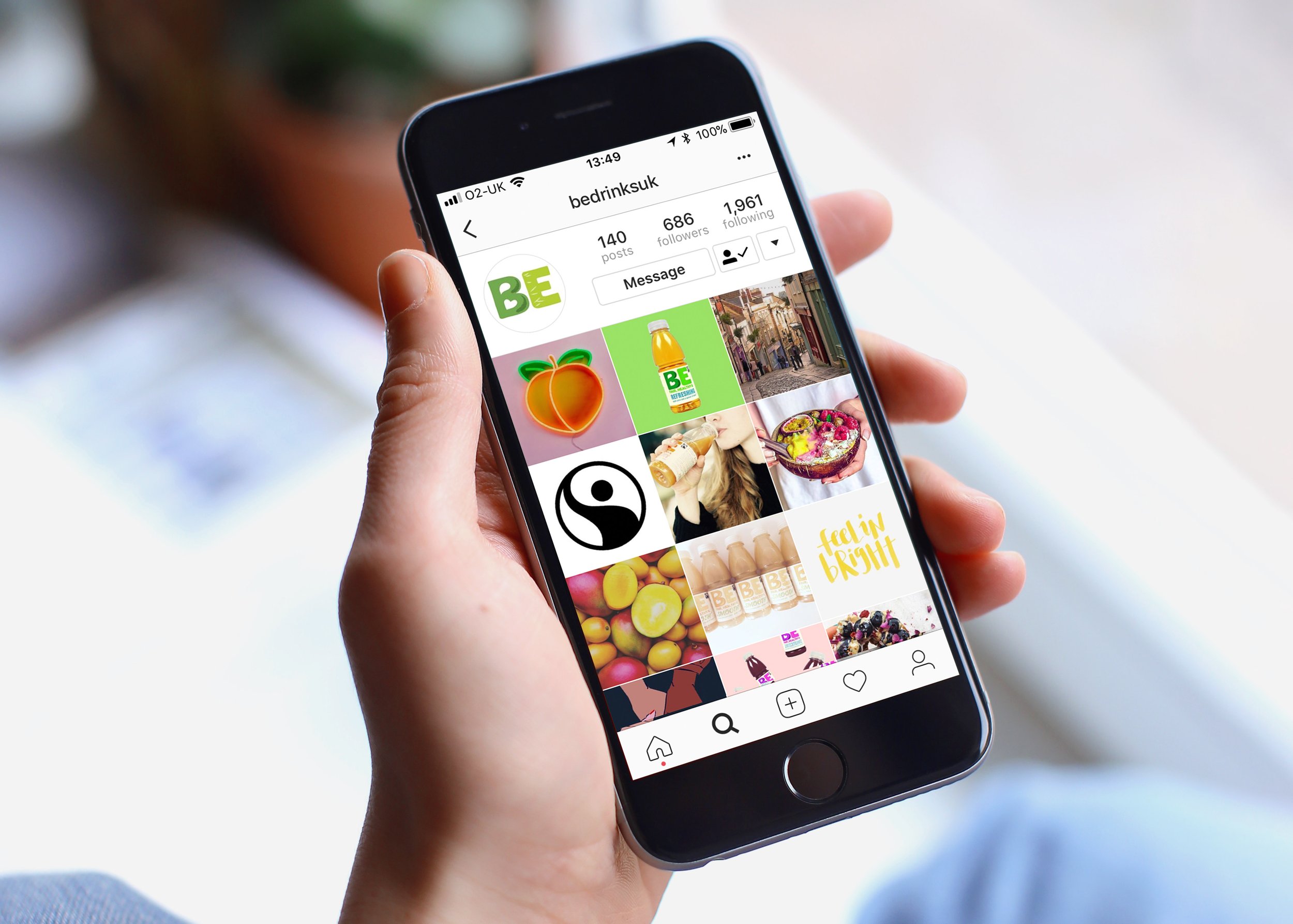 A person holding a smartphone displaying an Instagram profile page for BE Drinks with various images including food, drinks, street scenes, and icons.