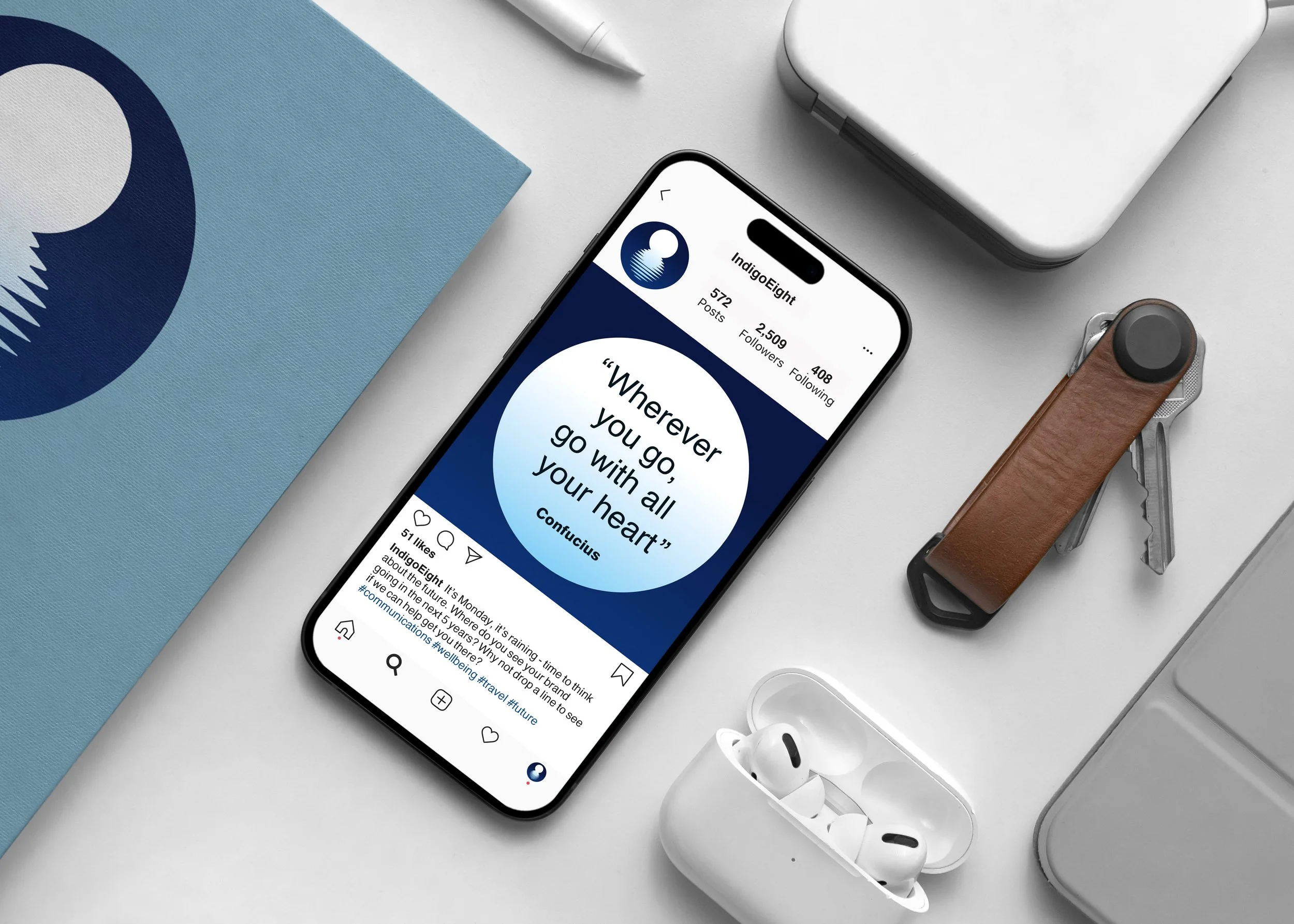 An iPhone displaying an Instagram profile of IndigoEight with a quote about the universe on the screen. The phone is on a white desk next to a pair of white wireless earbuds, a brown-handled keychain multitool, a blue notebook with a circular design, a white pen, and a closed white laptop.