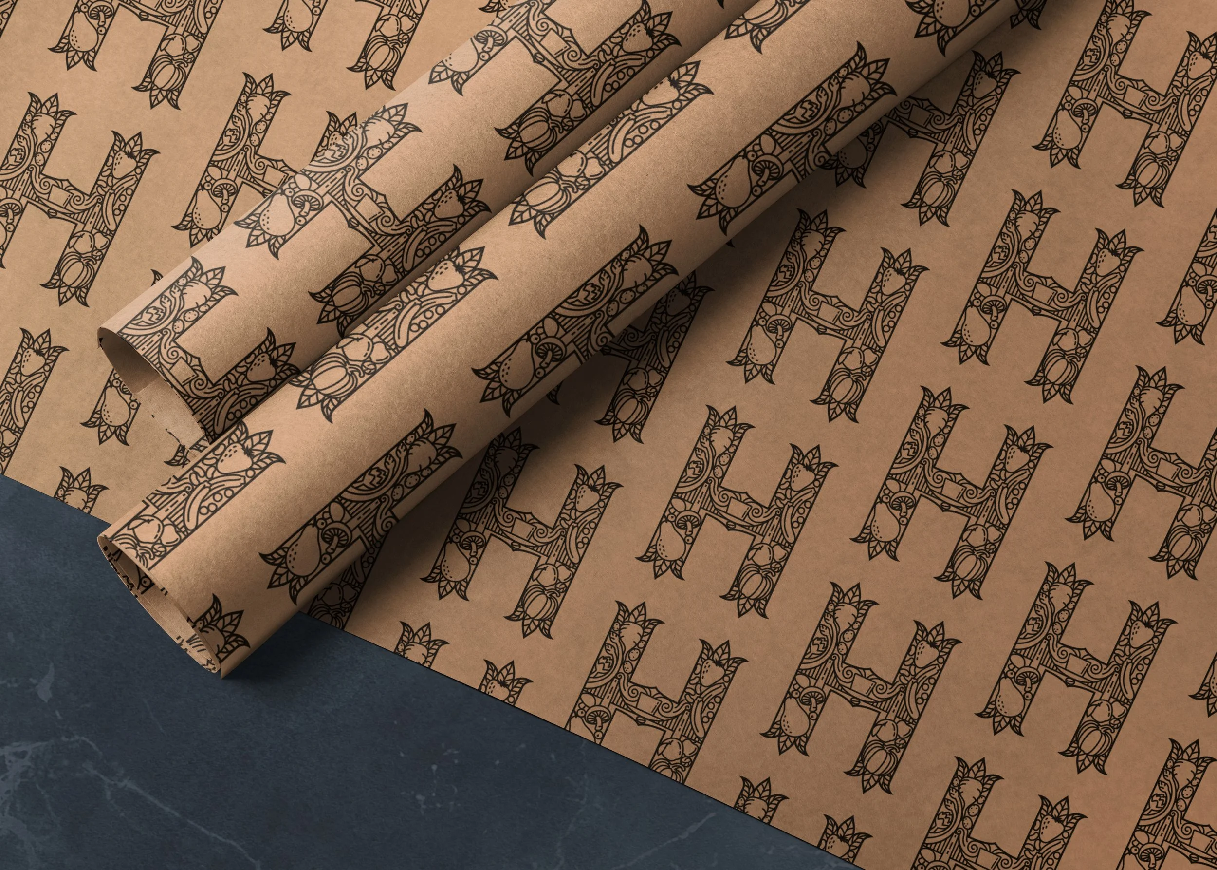 Wrapping paper with a pattern of ornate, stylised Hugo’s Greengrocer letter 'H' branding printed in black on brown kraft paper, partially unrolled on a dark surface.