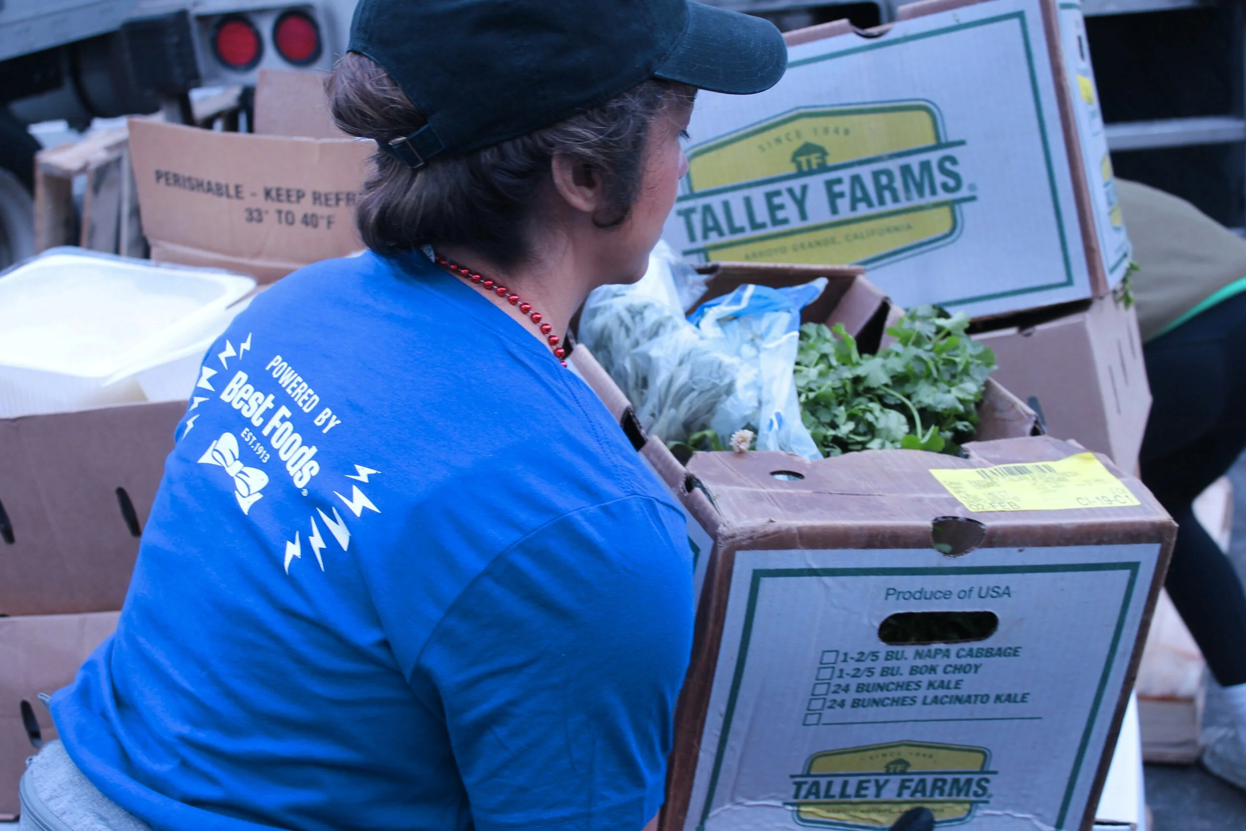 Person wearing a black cap and blue shirt with 'Powered by Best Foods' logo, next to boxes of fresh produce, including cabbage and kale, at a farmers market.