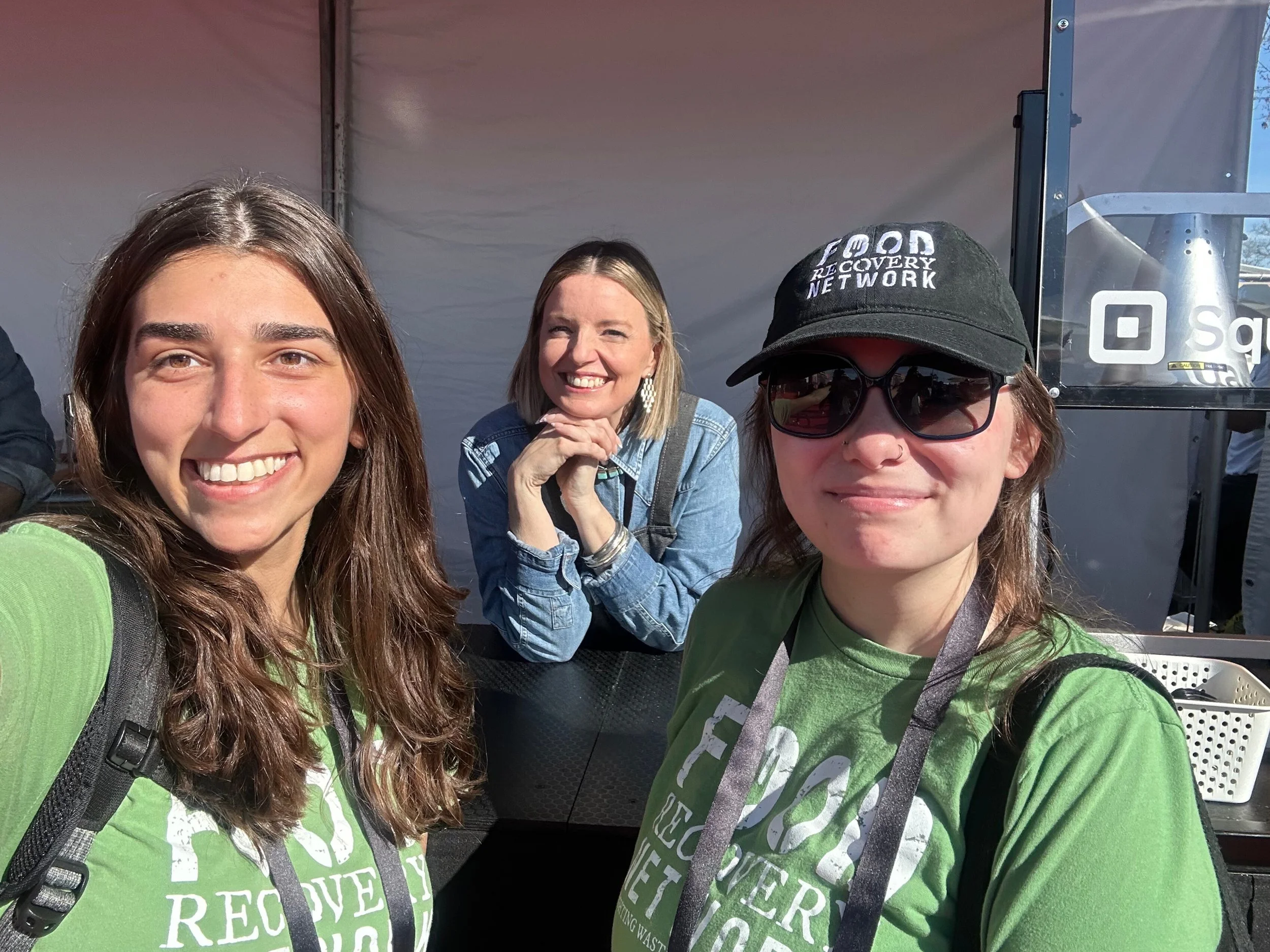 Three smiling women at an outdoor event, two wearing green shirts and one wearing a denim jacket and black cap with sunglasses.