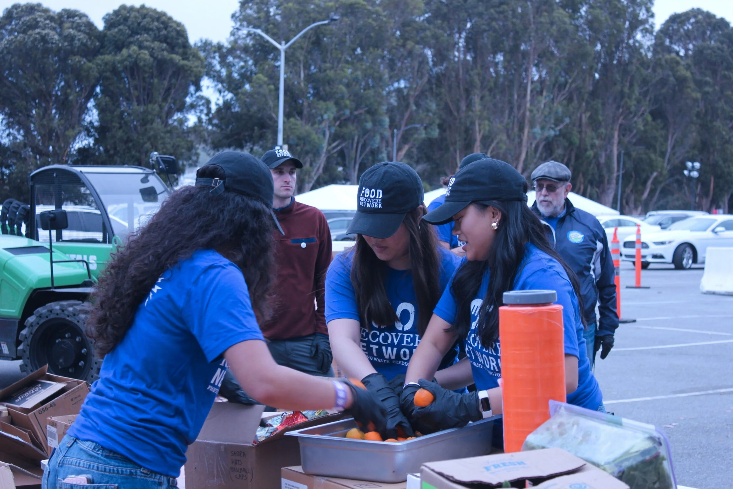 People in blue shirts and black hats packing food items in a parking lot during an outdoor event.