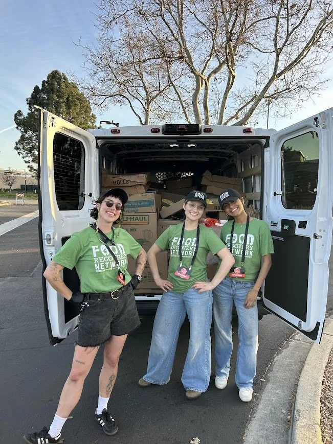 Three women wearing green T-shirts and black hats with 'Food Recovery Network' logos stand in front of an open white delivery van filled with boxes, with two trees and a clear sky in the background.