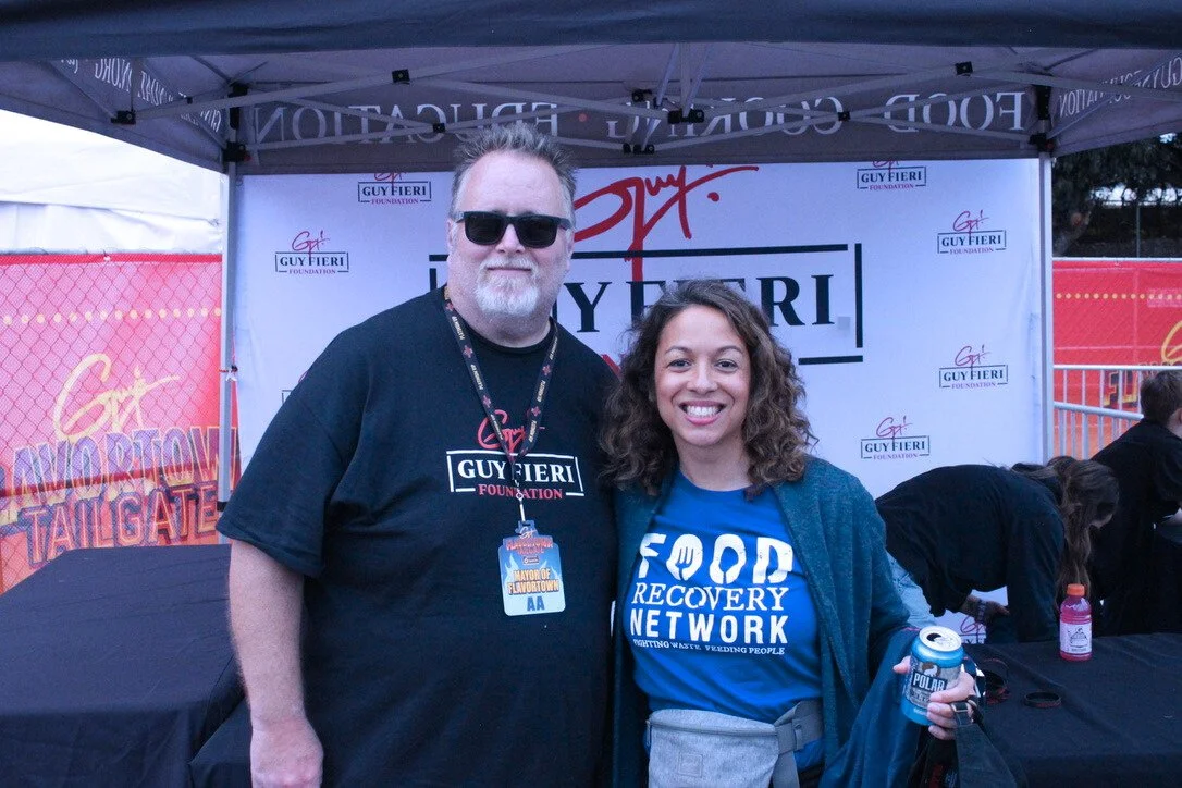 Two people standing in front of a Guy Fieri Foundation banner at an outdoor event, smiling for the camera.