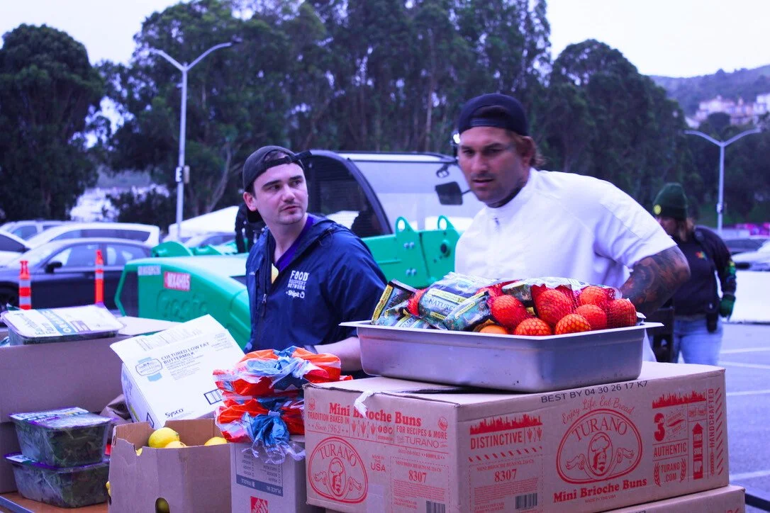 Two men standing behind a table filled with boxes of strawberries and other food items in a parking lot.