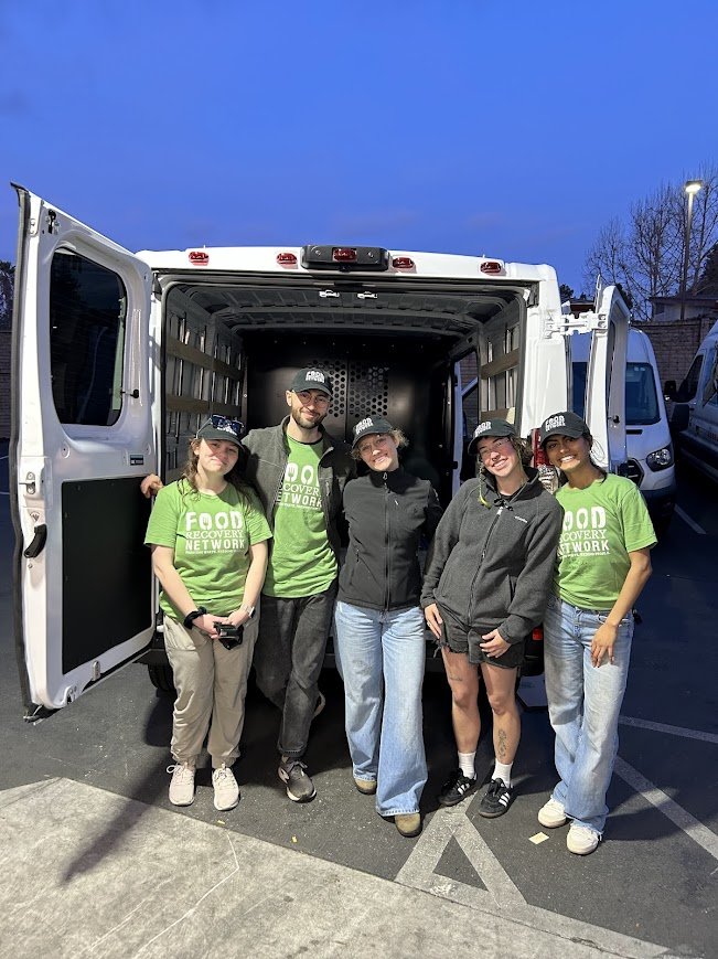 Five people standing in front of a white delivery van during dusk, with three women wearing green shirts that say 'Food Recovery Network' and two women in casual clothing, all smiling for the photo.