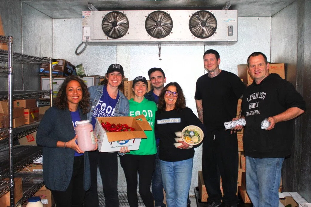 Group of seven people in a storage room holding food and supplies, smiling for the camera.