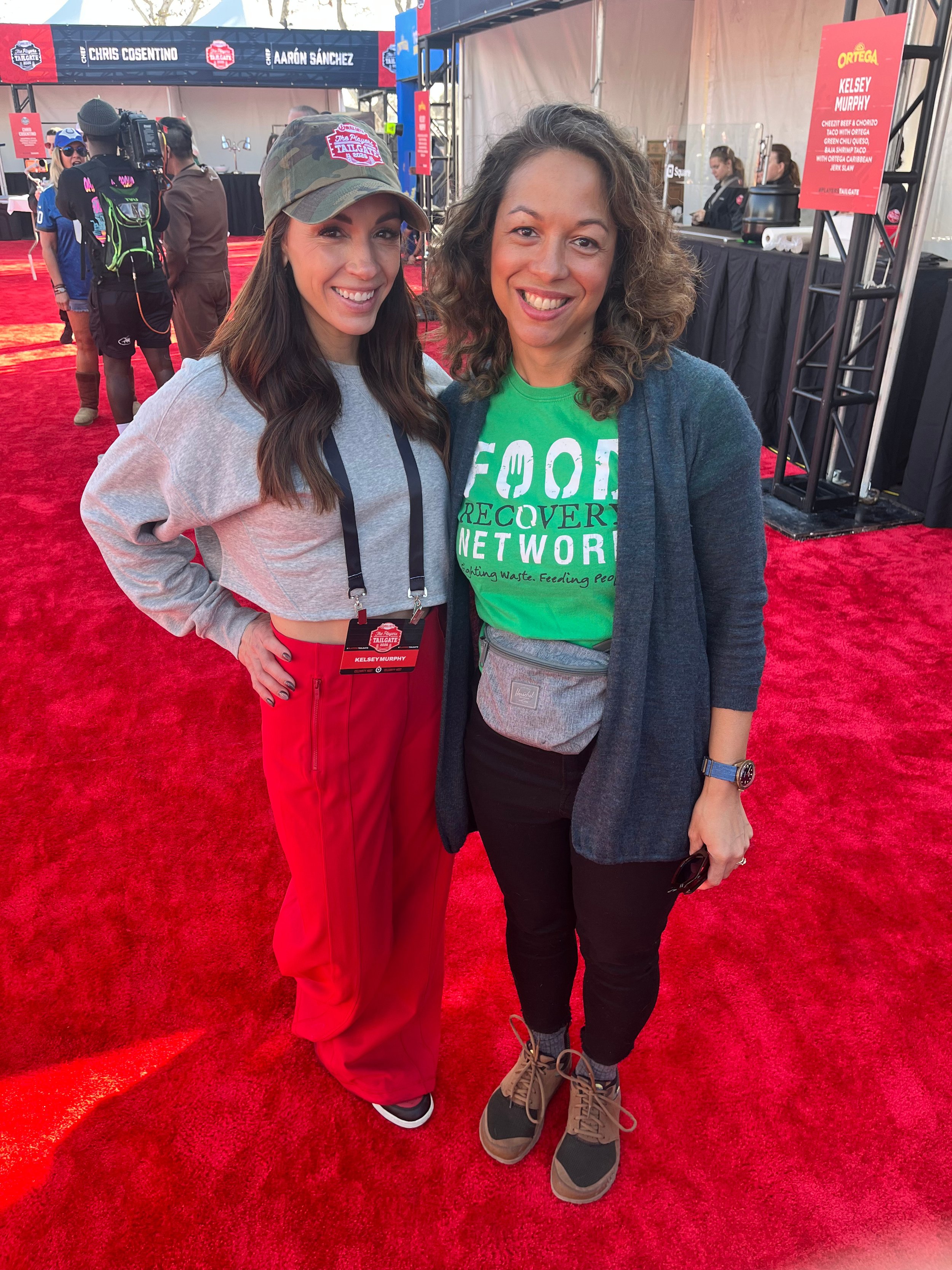 Two women smiling and standing close together at an outdoor event on a red carpet. The woman on the left has long brown hair, wearing a gray cropped sweatshirt and red pants, with a lanyard around her neck. The woman on the right has curly hair, wear