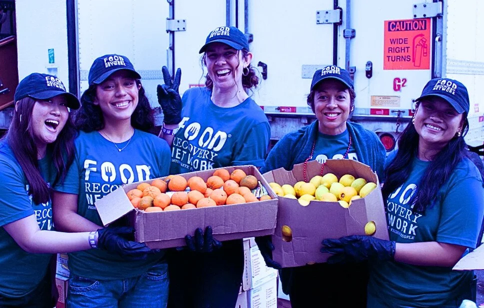 Five women wearing blue 'Food Recovery Network' T-shirts and caps, smiling and holding boxes of oranges and lemons at a food recovery event.
