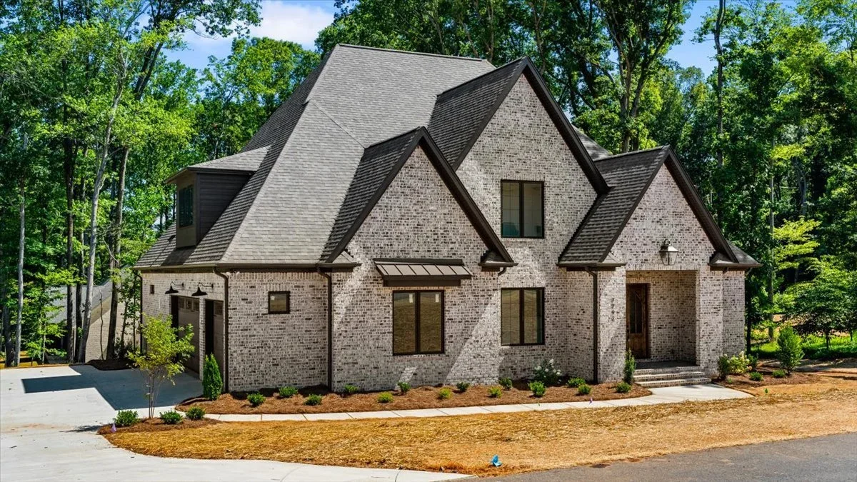 Brick house with steep rooflines, surrounded by trees, with a driveway and small front garden.