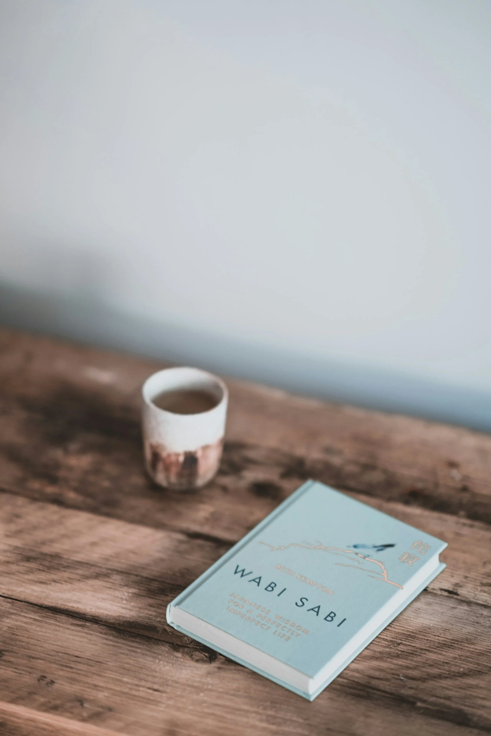 A wooden desk with a light blue book titled 'Wabi Sabi' by Waberi Sabii, a small ceramic cup, a blue notebook, and a corner of a paper or card.