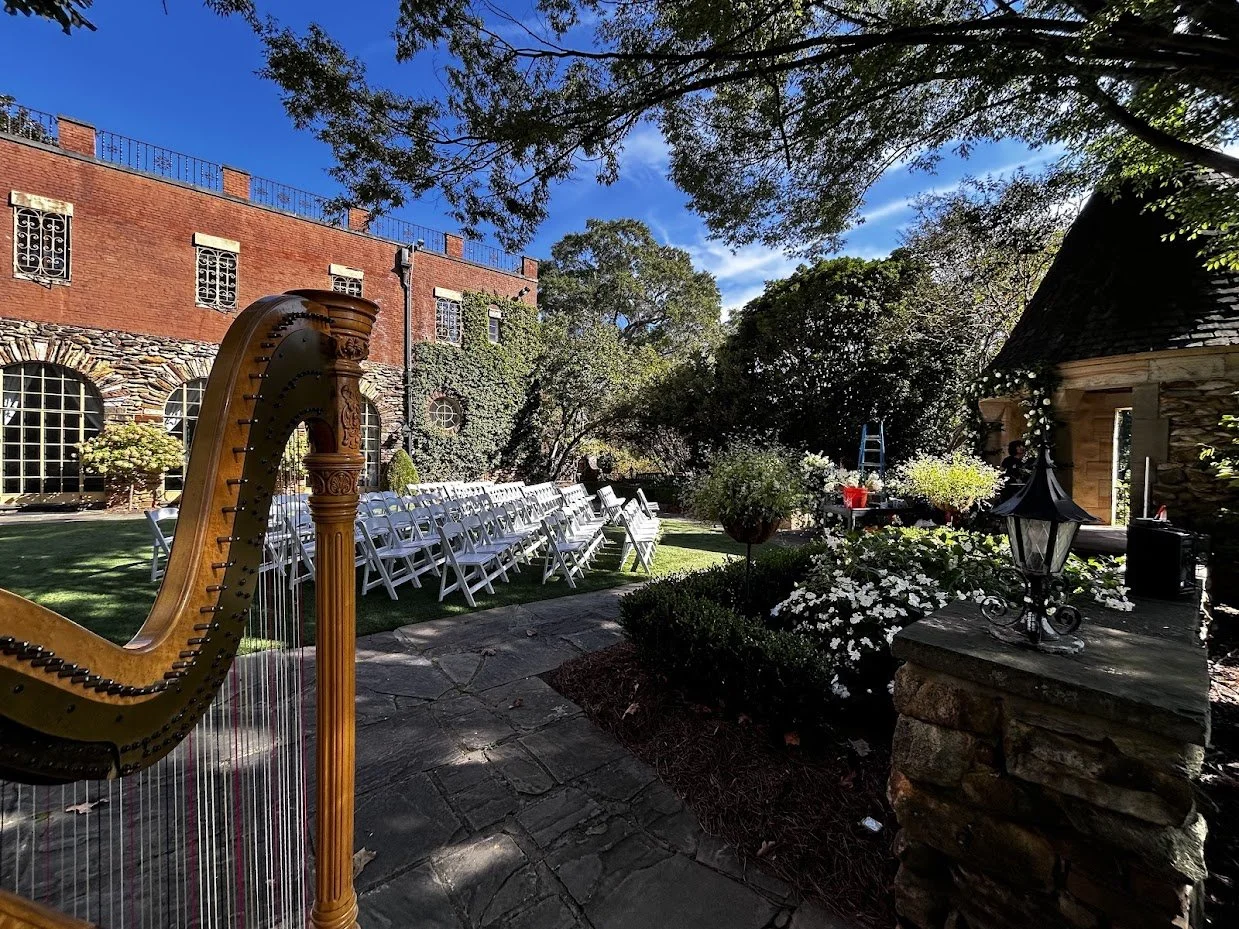 Outdoor garden patio with white chairs arranged in rows, a harp in the foreground, flowering bushes, and a brick building in the background under a blue sky.