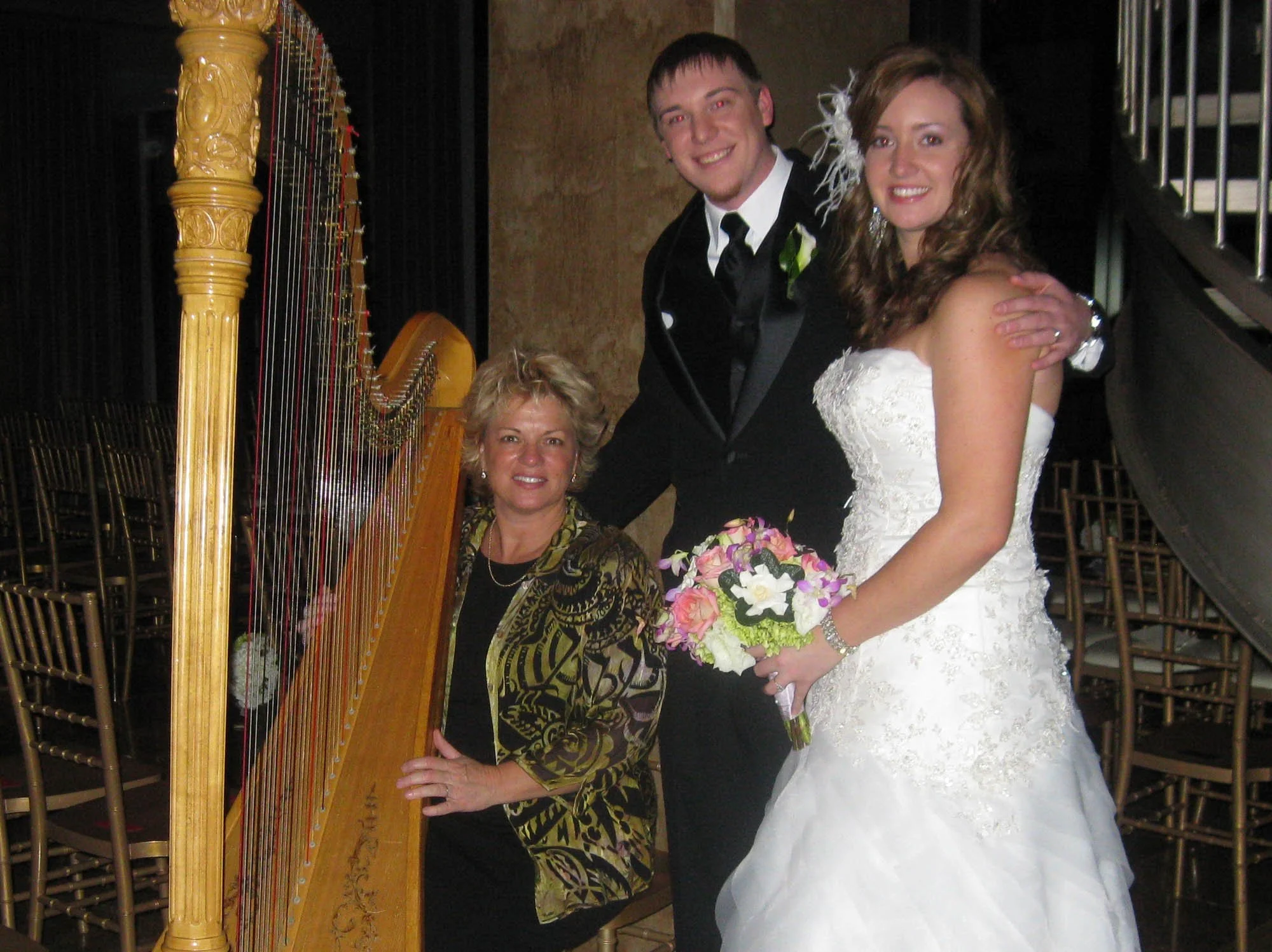 A bride and groom standing together at their wedding, with a woman playing a harp nearby. The bride is wearing a white lace wedding gown and holding a bouquet of flowers. The groom is dressed in a black tuxedo with a white shirt and black tie, and th