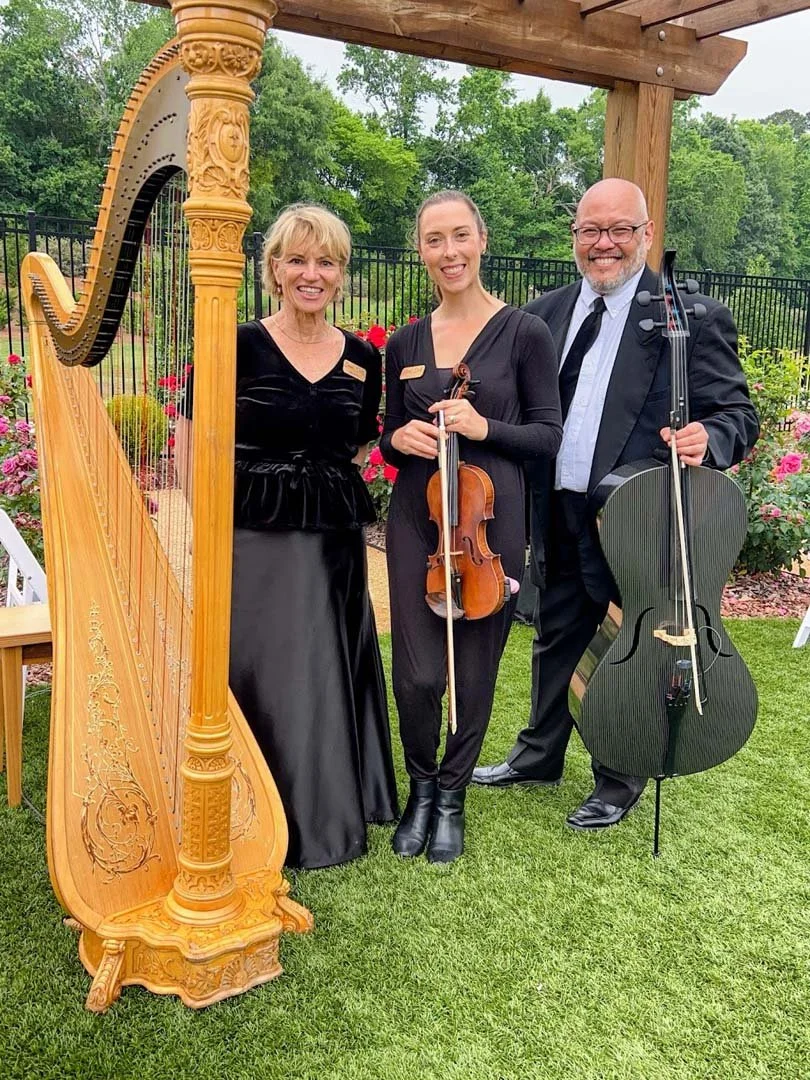 Three musicians, two women and one man, standing outdoors with string instruments, next to a large ornate harp. They are dressed in formal attire, smiling, with a garden and trees in the background.