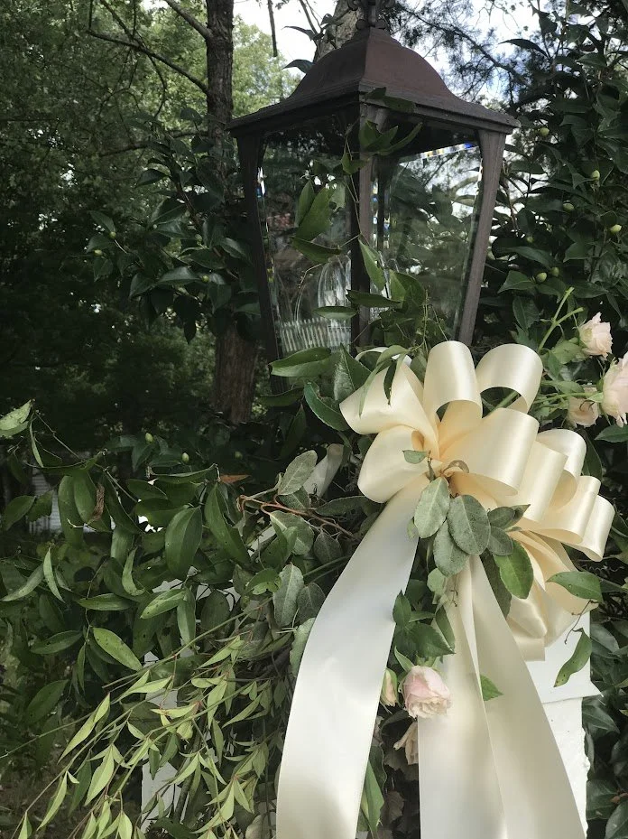 A decorative arrangement of greenery and pale pink flowers with a large cream-colored bow and ribbon, positioned near a black outdoor lantern in a garden setting.