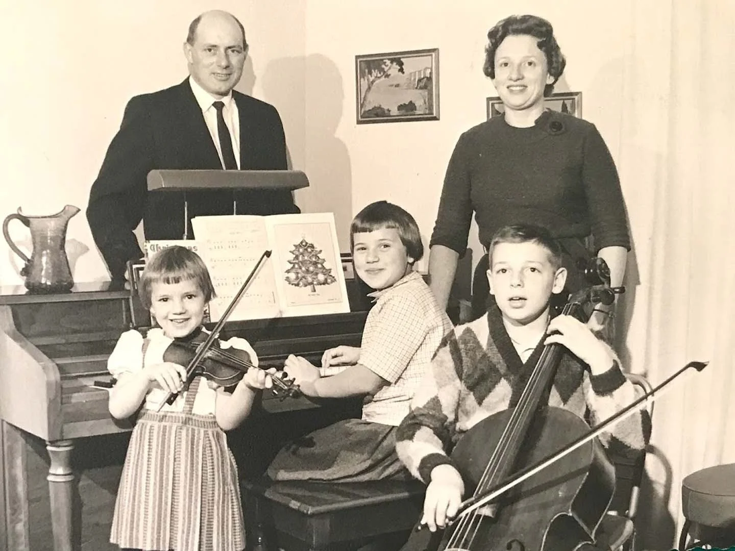 A black-and-white photo of a family in a room with musical instruments. A woman and two boys are seated at a piano, holding string instruments, while a man stands behind them. The woman is smiling, and the boys look surprised or curious. There are picture frames and a pitcher on a side table, with small paintings on the wall.