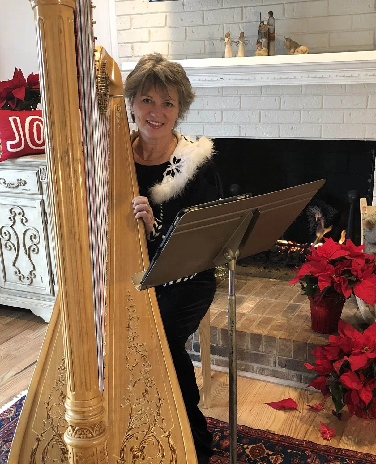 A woman with short, gray hair dressed in black, decorated with a white fur collar, playing a wooden harp in front of a fireplace with red poinsettia flowers nearby. She is smiling and looking at the camera, with a music stand and Christmas decoration