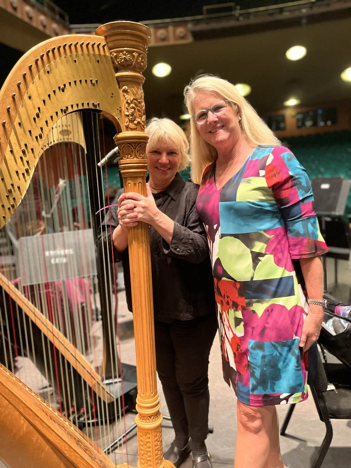 Two women smiling and posing with a harp in a concert hall.