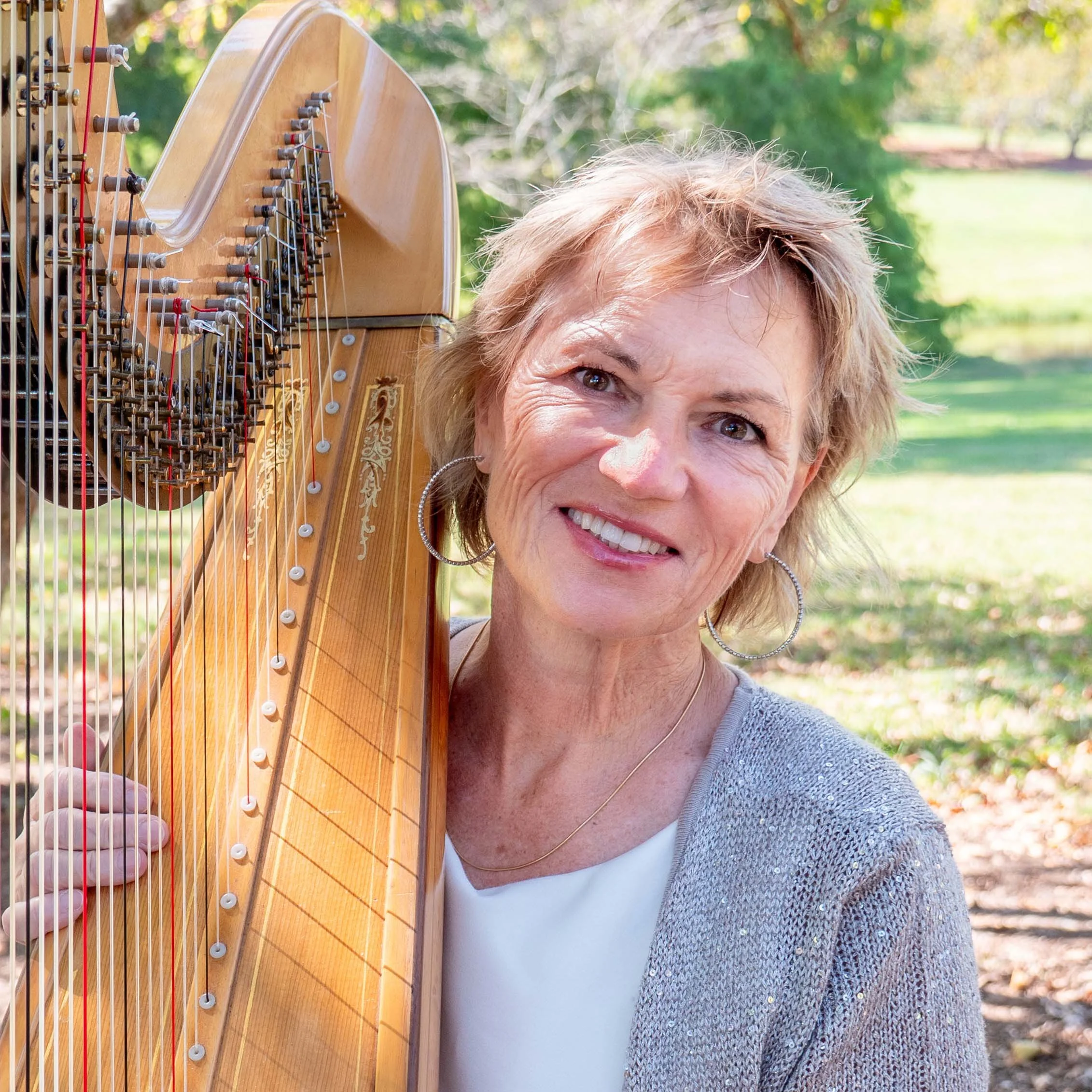 A woman smiling outdoors holding a harp in front of lush green trees.