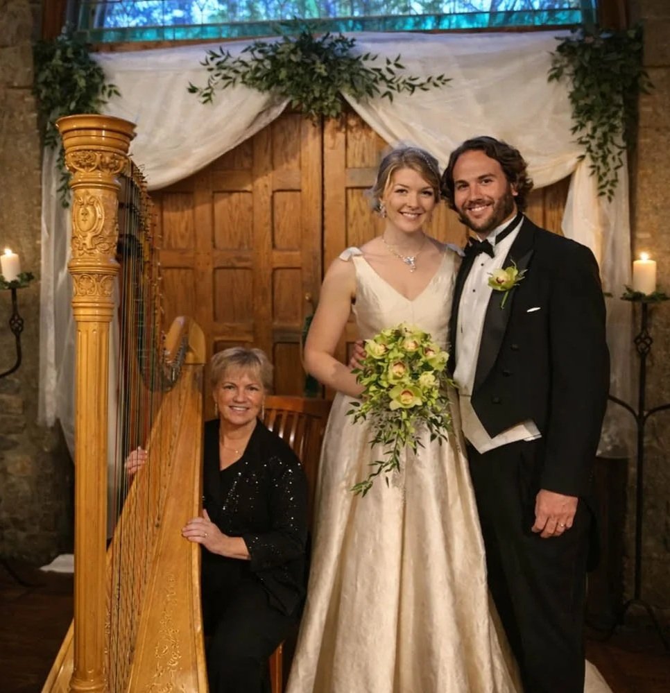 A bride and groom posing with a woman playing a harp at a wedding ceremony, decorated with draped fabric, candles, and greenery.