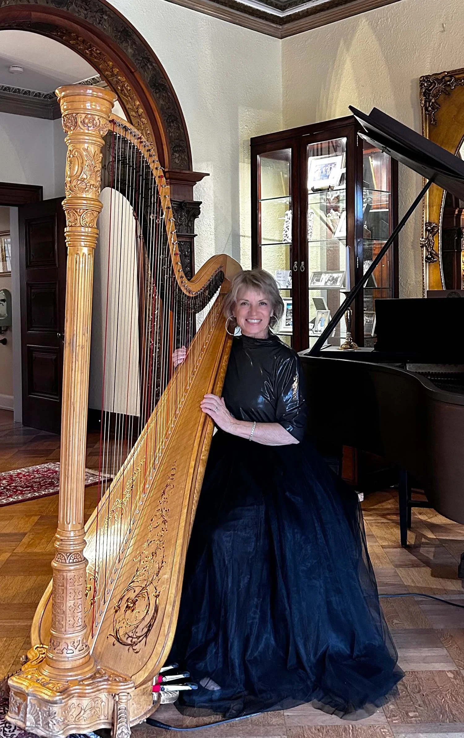 Woman in formal black dress smiling while playing a harp in a decorated room with a grand piano and display cabinet.