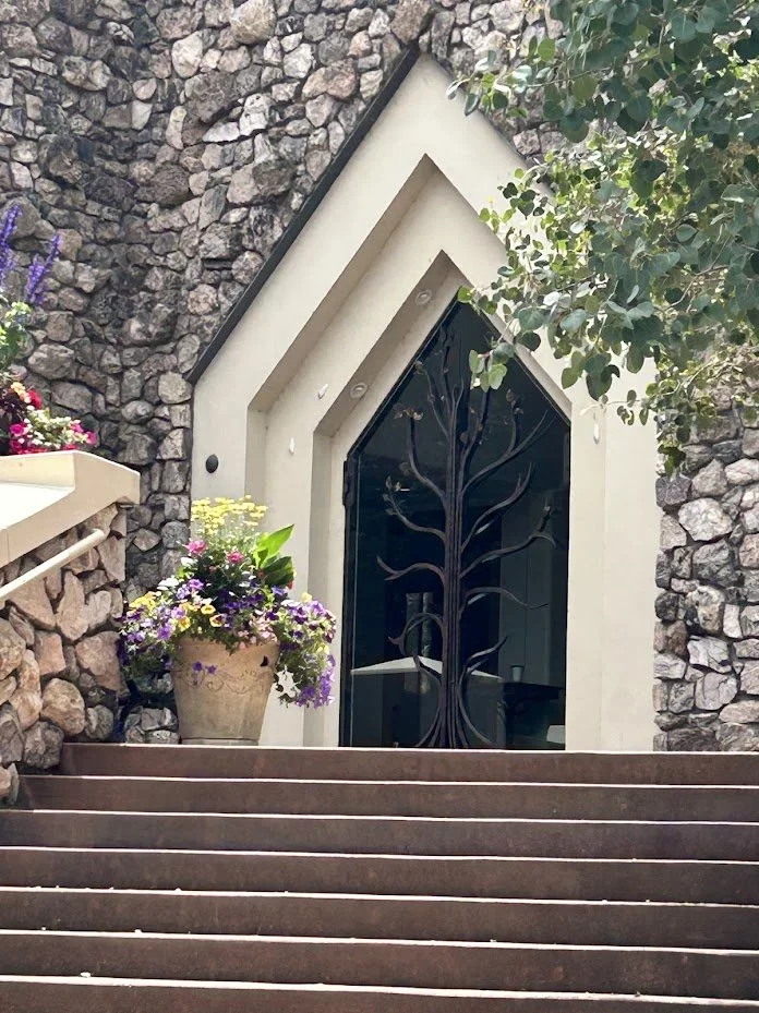 Stone building entrance with stairway, a black metal door featuring a tree design, and colorful potted flowers on the side.