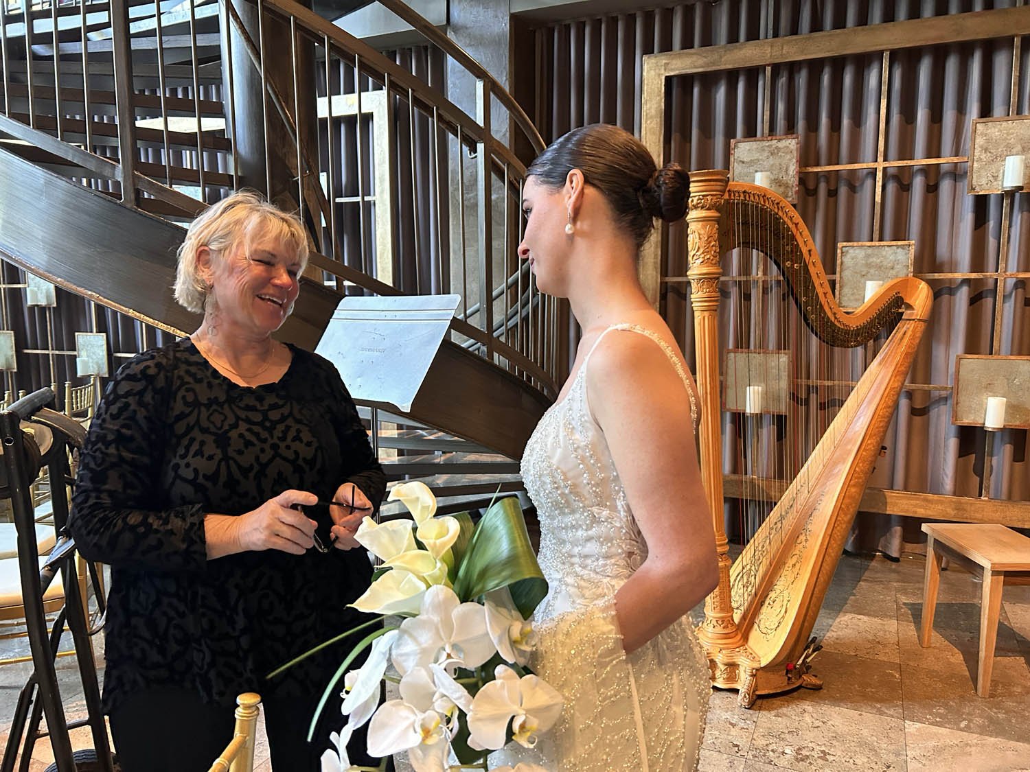 A woman in a wedding dress holding a bouquet of white flowers standing next to a harp, talking to another woman in black with blonde hair.