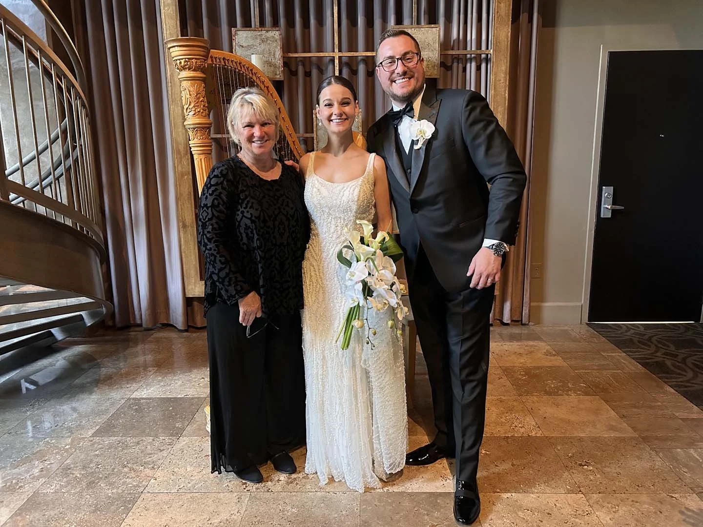 A bride in a white wedding dress holding a bouquet of white calla lilies stands between a woman in black and a man in a tuxedo, all smiling for a photo in an indoor venue with wood and stone decor.